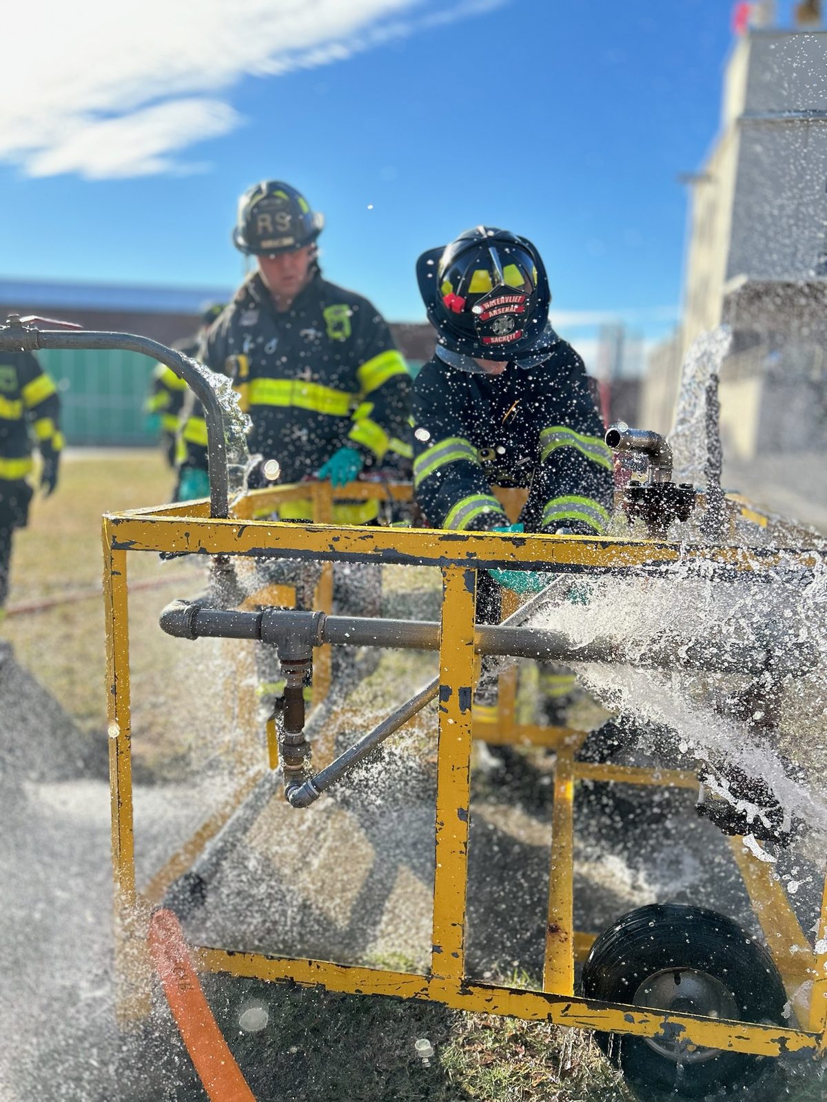 Firefighters in structural gear working a leaking pipe prop while water sprays across the frame.